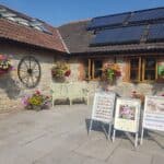 Courtyard Display Boards and Benches Bright courtyard with promotional display boards for food and cakes, hanging flower baskets, and a green bench in front of a stone building with rooftop solar panels.