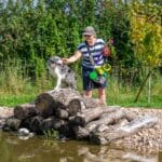Dog & Owner by Pond Woman in striped shirt interacts with a Border Collie perched on stacked logs beside a natural dog pond. She’s holding leashes, a ball launcher, and treat bags, under sunny skies.