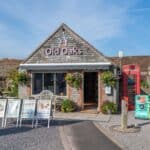 Old Oaks Reception and Shop (with boards & red phone box) Charming stone building marked "Old Oaks Reception & Shop", surrounded by hanging flower baskets, chalkboard signs, and food boards. A red British phone box and several picnic benches complete the welcoming scene.