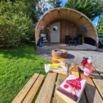 Sunny glamping setup with a wooden pod featuring a curved canvas canopy, outdoor seating, and a firepit. In the foreground, a wooden picnic table displays a book ("The State We’re In" by Adele Parks), snacks, cheeses, crackers, and bottled drinks. Surrounded by lush greenery and blue skies, the scene evokes a relaxed and leisurely atmosphere.