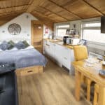 Open-plan holiday lodge interior featuring a cosy double bed with grey and green cushions, a black leather sofa, and a compact kitchen with white cabinetry, wooden worktops, and modern appliances. A wooden dining table set for two with wine and glassware sits beneath a wall-mounted TV. The space has a warm wooden ceiling and natural light from large windows, creating a bright and inviting atmosphere.