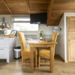 Compact and stylish holiday lodge kitchen and dining area featuring a wooden table set for two with mustard yellow upholstered chairs, a bottle of wine, and glassware. The kitchenette includes a microwave, toaster, and air fryer on wooden countertops. A flat-screen TV is mounted under a sloped wooden ceiling, and natural light streams in through large windows.