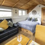 Cosy lodge interior featuring a double bed with grey bedding and green and blue scatter cushions, a black leather sofa with yellow and green cushions, and a sloped wooden ceiling. The space includes wooden flooring, soft lighting, and windows letting in natural light, creating a warm and welcoming atmosphere.