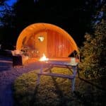 Tranquil nighttime glamping scene featuring a wooden pod with a curved canvas roof, softly illuminated by the glow of a firepit. A wicker chair with a blanket, a picnic bench with a lantern, and warm lighting create a cosy, peaceful atmosphere. Surrounded by trees and greenery, with a car parked nearby on the gravel.