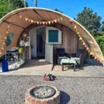Quirky glamping pod with a curved canvas awning decorated with red and yellow bunting, set on a gravel pitch with a wooden deck. The setup includes outdoor seating with a checkered tablecloth, a portable kitchen area with cooking supplies, and a firepit in the foreground. Surrounded by greenery, under a clear blue sky, creating a fun and relaxed camping atmosphere.