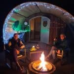 Cosy glamping evening scene with two people sitting around a lit firepit outside a wooden pod shelter, decorated with fairy lights and colourful bunting. The guests are wrapped up warm, smiling with drinks in hand, while candles and soft lighting create a warm, festive glow against the dark night.