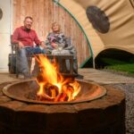 Smiling couple sitting outside a wooden glamping pod under a canvas awning, enjoying drinks by a glowing firepit. Wrapped in a cosy blanket, they’re seated on a decked area with a rustic wooden backdrop, as the flames provide warmth and a welcoming glow against the night.